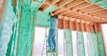 Male builder insulating wooden frame house. Man worker spraying polyurethane foam inside of future cottage, using plural component gun. Construction and insulation concept.